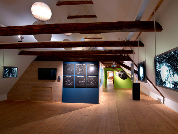 Blick in die Ausstellung in einem ausgebauten Dachboden eines alten Bauernhauses. Von der Decke hängen Planetenattrapen, an der Seite sind beleuchtete Fotografien aus dem Weltraum zu sehen.  