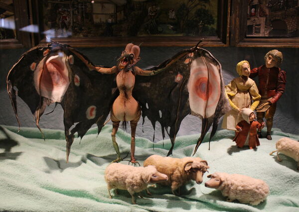 Blick in eine Vitrine mit Puppen aus einem Animationsfilm. Ein teuflisches Fabelwesen breitet die Flügel aus. Dahinter steht eine Familie, im Vordergrund weidet eine Gruppe Schafe.