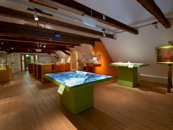 Blick in die Ausstellung im ausgebauten Dachstuhl eines alten bauernhauses. In der Mitte stehen Vitrinen und ein Tisch mit einer Landkarte. 