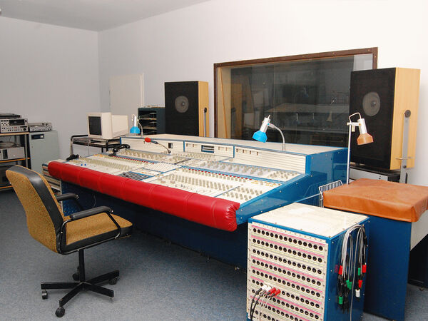 Im Tonstudio steht ein altes Mischpult vor einer Scheibe, die zur Aufnahmekabine gehört.