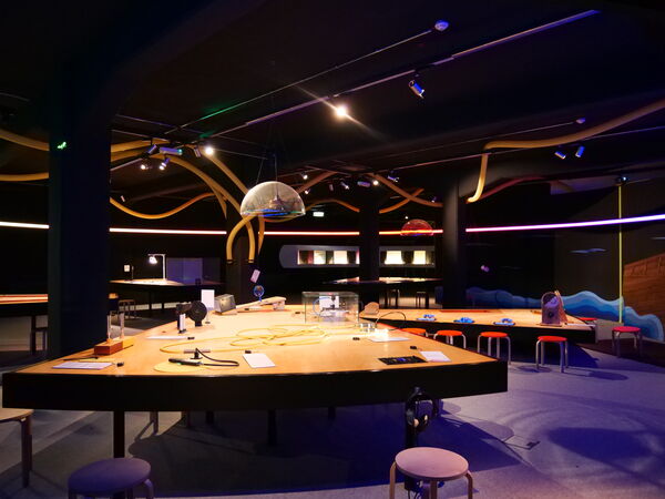 Blick in den Ausstellungsraum. In der Mitte stehen große Tische mit interaktiven Exponaten.